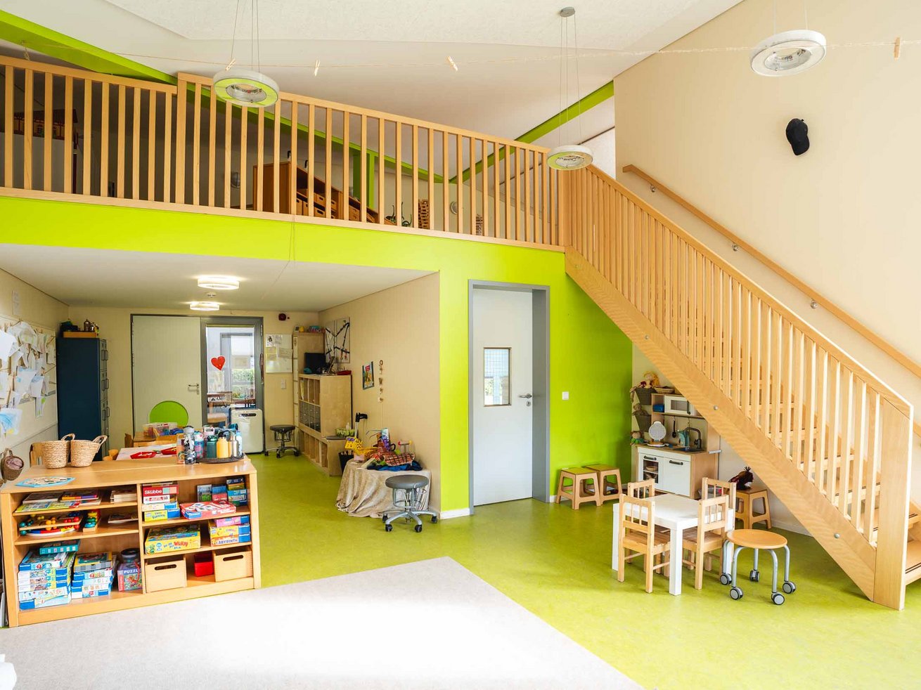 Heller Gruppenraum der Kita mit Spielecke, Kinderküche, Tischen und Treppe zur oberen Ebene.