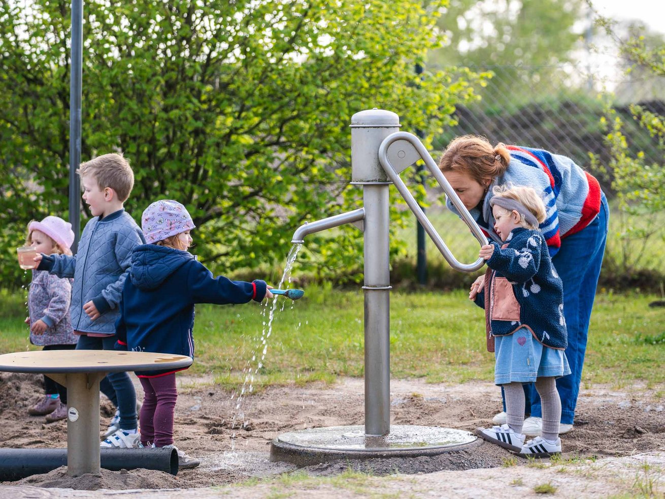 Kinder spielen gemeinsam mit einer Erzieherin an einer Wasserpumpe im Außengelände der Kita.