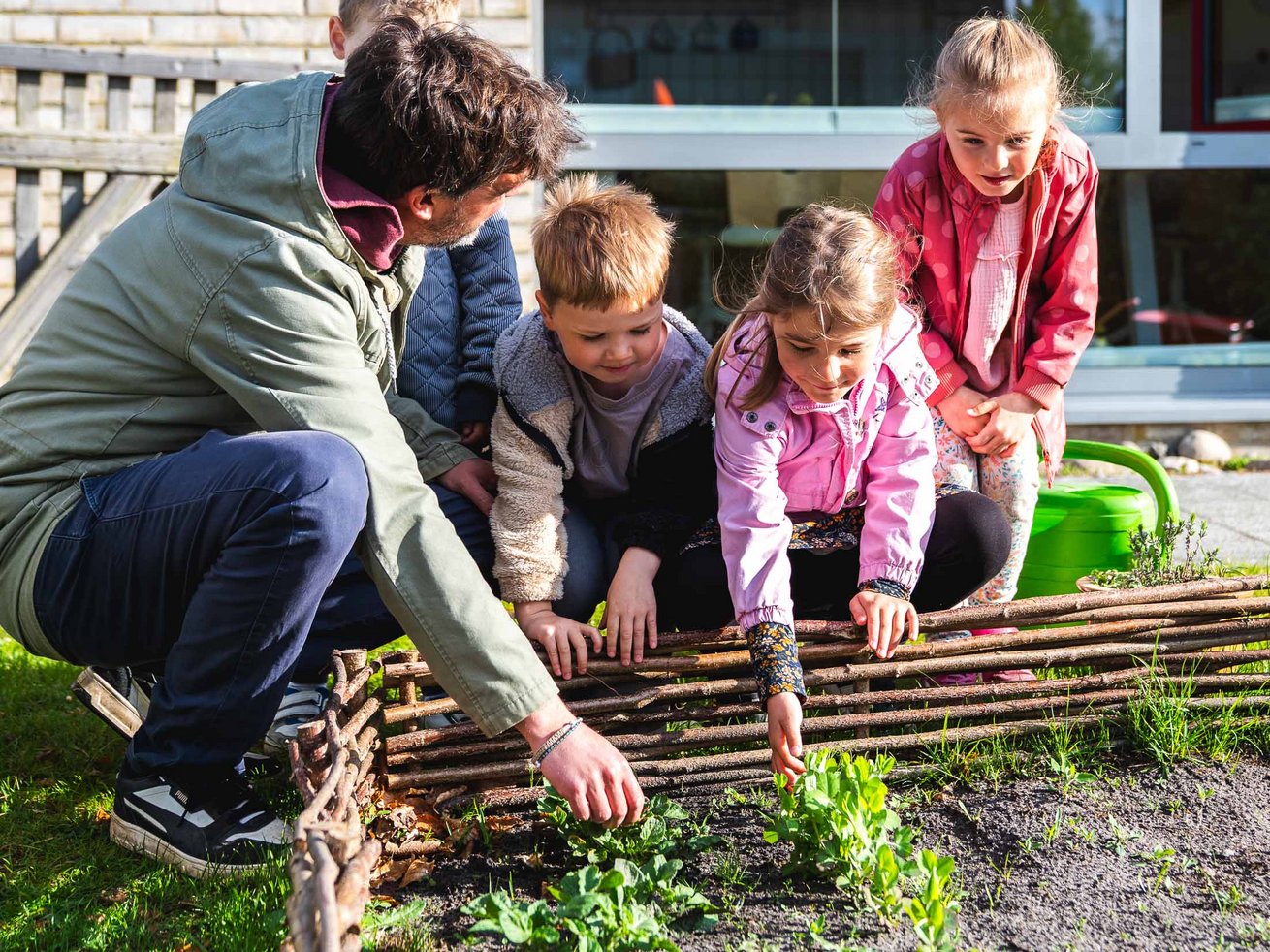 Kinder entdecken gemeinsam mit einem Erzieher Pflanzen in einem Beet im Garten der Kita.